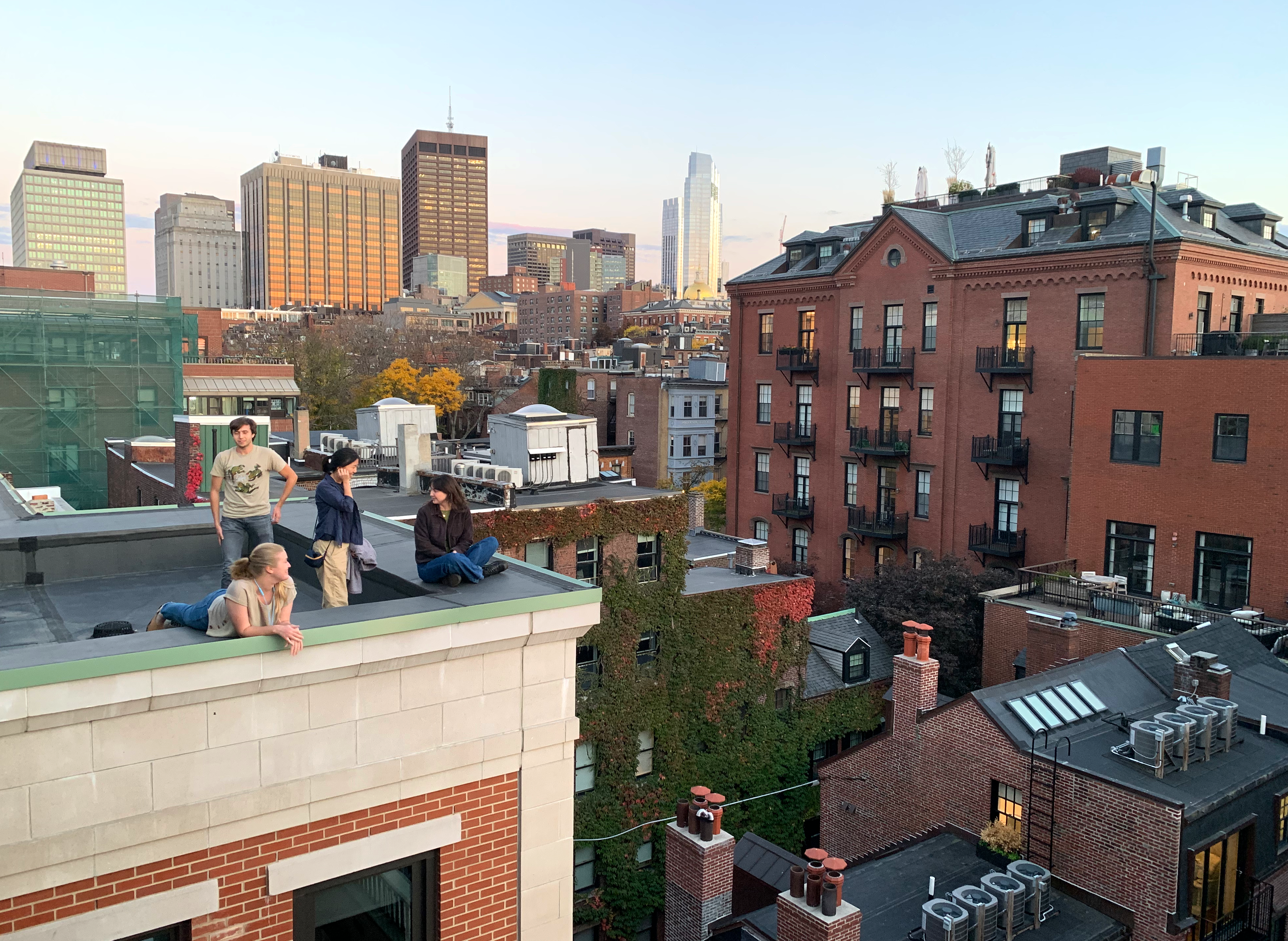 Four people sit (safely) near the edge of a roof. They are surrounded by Beacon Hill’s rooftops and a hint of a sunset.